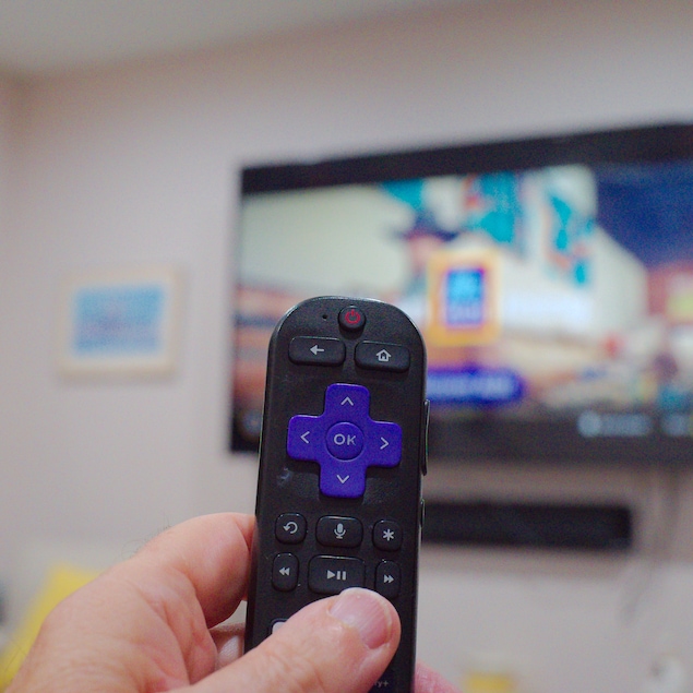 Une main tenant une télécommande Roku devant une télévision.