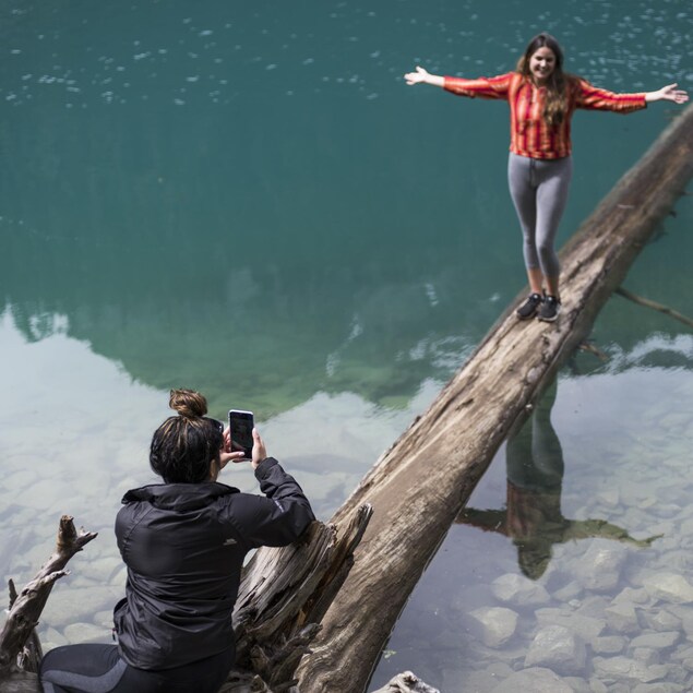 Une jeune femme prend son amie en photo sur un tronc à demi-submergé aux abords d'un lac du parc britanno-colombien de Joffre Lakes.