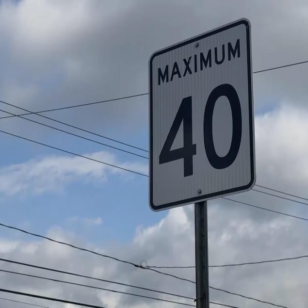 Un panneau de signalisation routière rectangulaire blanc, bordé de noir, affiche l'inscription « MAXIMUM 40 » en lettres et chiffres noirs. Il est fixé sur un poteau métallique face à un ciel nuageux traversé par plusieurs fils électriques.