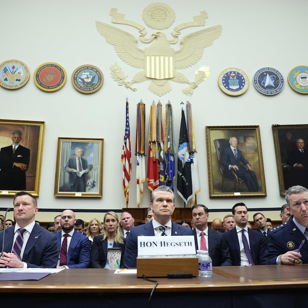Trois hommes en veston ou uniforme militaire sont assis à une longue table devant des sceaux officiels et des portraits