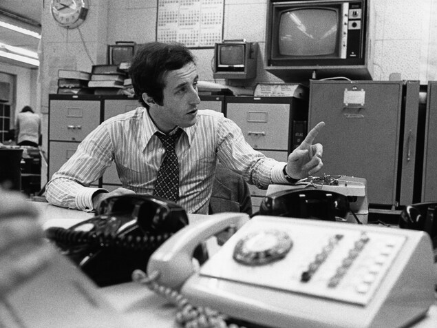 Dans une salle de nouvelles, l'animateur Bernard Derome, est assis à un bureau sur lequel il y a plusieurs appareils de téléphone. À l'arrière-plan, on voit des classeurs appuyés au mur où des téléviseurs sont accrochés. Dans une salle de nouvelles, l'animateur Bernard Derome, est assis à un bureau sur lequel il y a plusieurs appareils de téléphone. À l'arrière-plan, on voit des classeurs appuyés au mur où des téléviseurs sont accrochés.