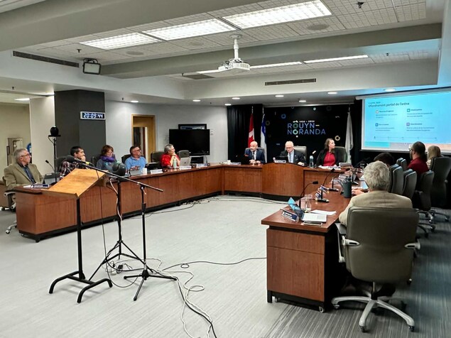 Les conseillers et conseillères municipaux de Rouyn-Noranda regarde un texte afficher sur un toile sur l'effondrement partiel de l'aréna d'Évain.