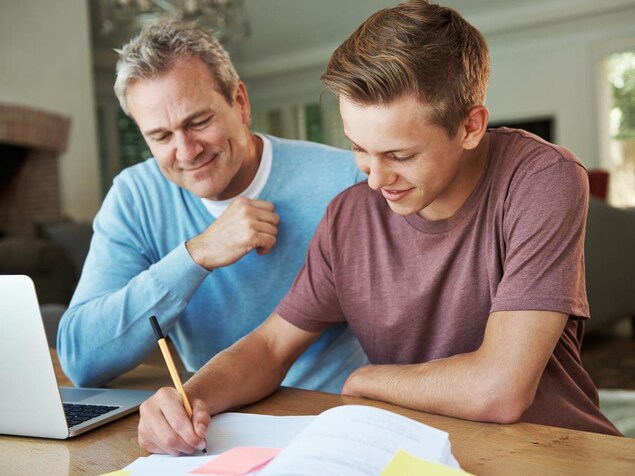 Un adolescent écrit dans un cahier, son père à ses côtés. Un adolescent écrit dans un cahier, son père à ses côtés.