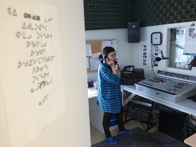 Mary Koperqualuk parle au téléphone devant une console radio. Mary Koperqualuk parle au téléphone devant une console radio.