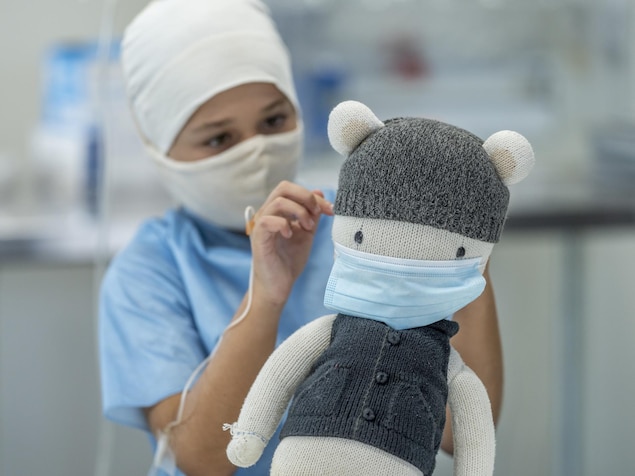 Un enfant de 9 ans assis dans un hôpital avec une perfusion, un masque et un bonnet.