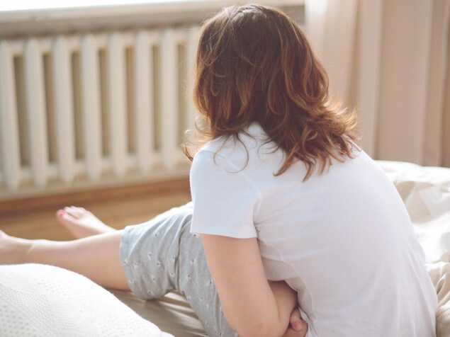 Une femme vue de dos, assise sur son lit, les bras repliés sur son ventre. Une femme vue de dos, assise sur son lit, les bras repliés sur son ventre.