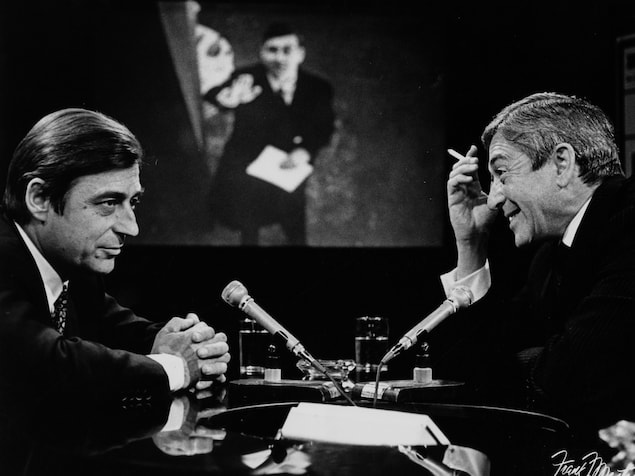 Dans un studio de télévision, François Périer s'entretient avec Fernand Seguin. Une image floue de François Périer, probablement dans un de ses films, est projetée sur le mur du fond. Dans un studio de télévision, François Périer s'entretient avec Fernand Seguin. Une image floue de François Périer, probablement dans un de ses films, est projetée sur le mur du fond.