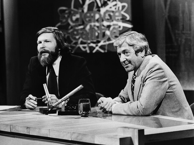 Dans un studio de télévision, Joël Le Bigot et Fernand Seguin assis à une table surmontée de deux microphones. En arrière-plan, on voit le titre de l'émission sur un écran au mur Dans un studio de télévision, Joël Le Bigot et Fernand Seguin assis à une table surmontée de deux microphones. En arrière-plan, on voit le titre de l'émission sur un écran au mur
