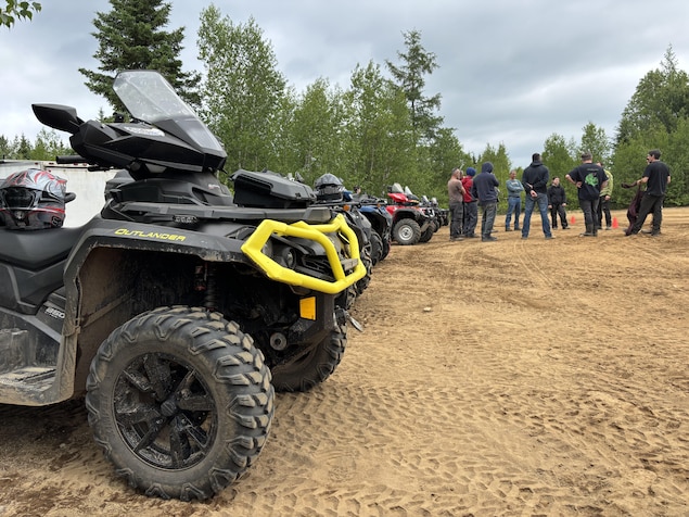 Sur un terrain sablonneux, quelques personnes sont réunies en cercle au loin. Tout près, des véhicules tout-terrains sont stationnés en rangée. 