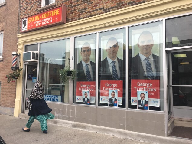 Des affiches sont installées dans la vitrine du local électoral du candidat Tsantrizos. À côté, une enseigne annonce un salon de coiffure pour homme. Sur la rue marche une femme qui porte un voile. Des affiches sont installées dans la vitrine du local électoral du candidat Tsantrizos. À côté, une enseigne annonce un salon de coiffure pour homme. Sur la rue marche une femme qui porte un voile.