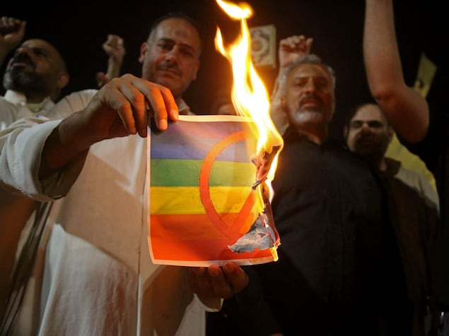 Une personne entourée d'autres personnes tient une feuille en train de brûler avec le drapeau LGBTQ+ avec un signe rouge d'interdiction dessus.