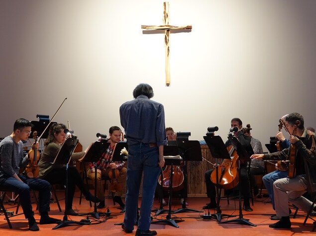 Le maestro debout, devant les musiciens. Sur le mur, une croix. Le maestro debout, devant les musiciens. Sur le mur, une croix.