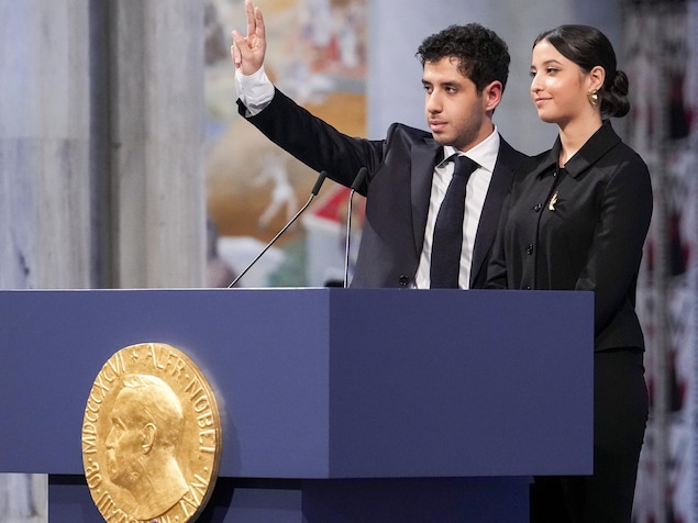 Une jeune femme et un jeune homme, qui fait le signe de la paix de sa main droite, sourient pensivement, debout derrière un lutrin.