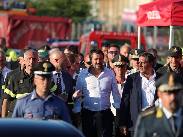 Un homme, entouré de pompiers ou policiers ainsi que d'hommes en veston-cravate, marche en regardant droit devant lui. Le soleil semble se coucher dans cette photo. Un homme, entouré de pompiers ou policiers ainsi que d'hommes en veston-cravate, marche en regardant droit devant lui. Le soleil semble se coucher dans cette photo.