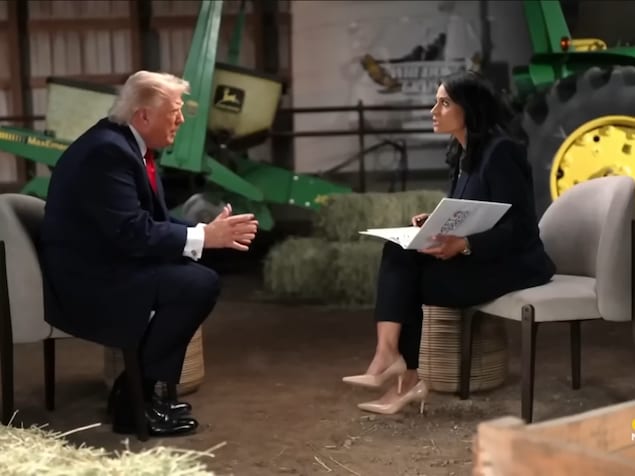 Un homme est assis au bout de sa chaise en gesticulant, une femme parle avec lui. Des tracteurs et des bottes de foin sont disposés en arrière-plan.
