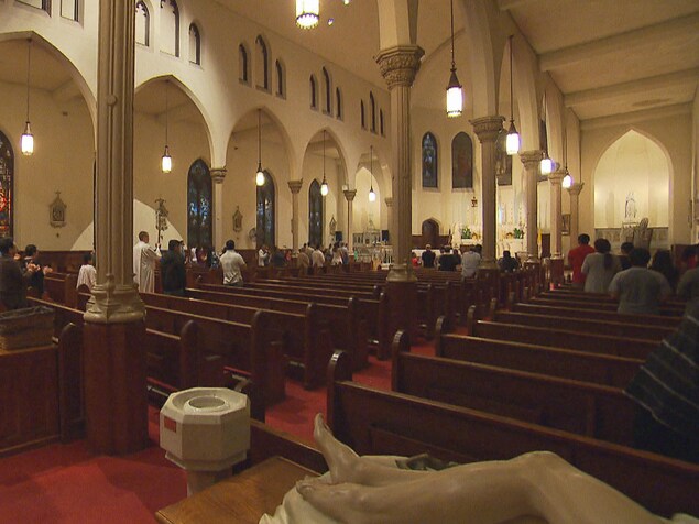 Des personnes se tiennent debout dans une église, alors qu'il semble faire sombre à l'extérieur. Elles sont peu nombreuses. Des personnes se tiennent debout dans une église, alors qu'il semble faire sombre à l'extérieur. Elles sont peu nombreuses.