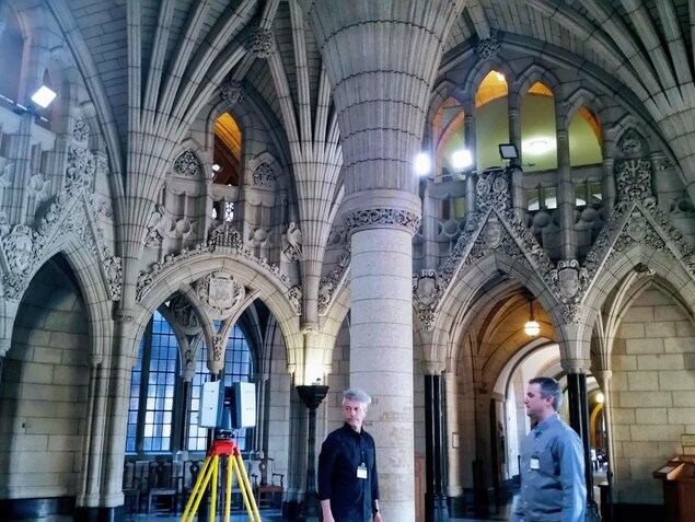 On voit deux hommes dans l'édifice du centre du parlement d'Ottawa, à côté d'eux se trouve un appareil de mesure sur trépied : autour d'eux, on voit les colonnes, les arches, les alcôves. On voit deux hommes dans l'édifice du centre du parlement d'Ottawa, à côté d'eux se trouve un appareil de mesure sur trépied : autour d'eux, on voit les colonnes, les arches, les alcôves.