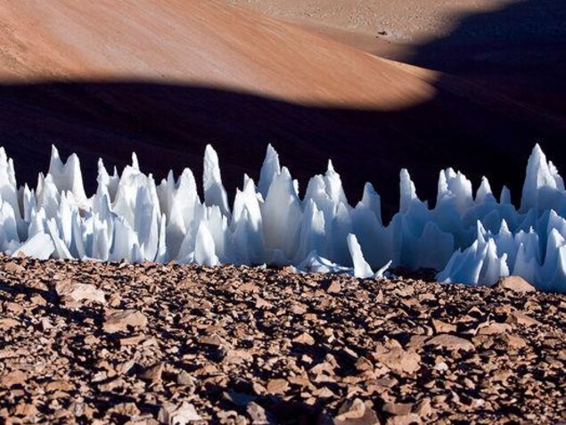 On voit des formations de glace en forme de pics, formant une bande horizontale, au sol. Le reste du terrain est rocheux. On voit des formations de glace en forme de pics, formant une bande horizontale, au sol. Le reste du terrain est rocheux.