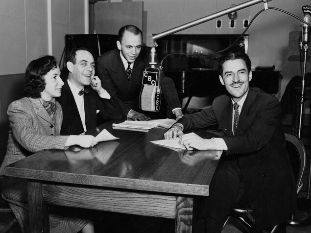 Dans un studio de radio, Lisette LeRoyer, Miville Couture, Roger De Vaudreuil et René Lecavalier, sont assis autour d'une table, un micro suspendu entre eux. Dans un studio de radio, Lisette LeRoyer, Miville Couture, Roger De Vaudreuil et René Lecavalier, sont assis autour d'une table, un micro suspendu entre eux.