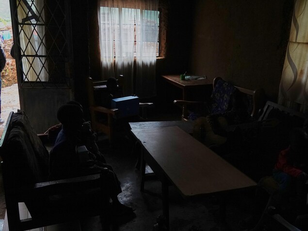 Salon à l'intérieur d'une maison avec deux fenêtres drapées de rideaux et une porte ouverte donnant sur l'extérieur. Des enfants sont assis dans l'obscurité autour d'une table. Salon à l'intérieur d'une maison avec deux fenêtres drapées de rideaux et une porte ouverte donnant sur l'extérieur. Des enfants sont assis dans l'obscurité autour d'une table.