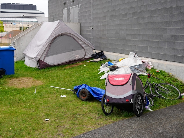 Une tente grise et des effets personnels sont installés sur le gazon au pied d'un mur en béton.