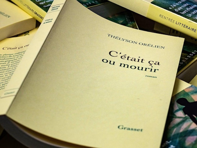 A copy of Orélien's novel "C'était ça ou mourir", open and face down, on a pile of other copies of the same book.