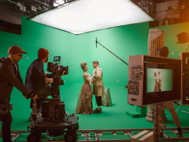 Une équipe tourne une scène de film dans un studio devant un écran vert, avec un acteur et une actrice habillés en costumes d'époque au centre. 