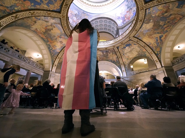 Un femme portant un drapeau sur ses épaules est debout à l'arrière d'une salle rempli de personnes assises. 