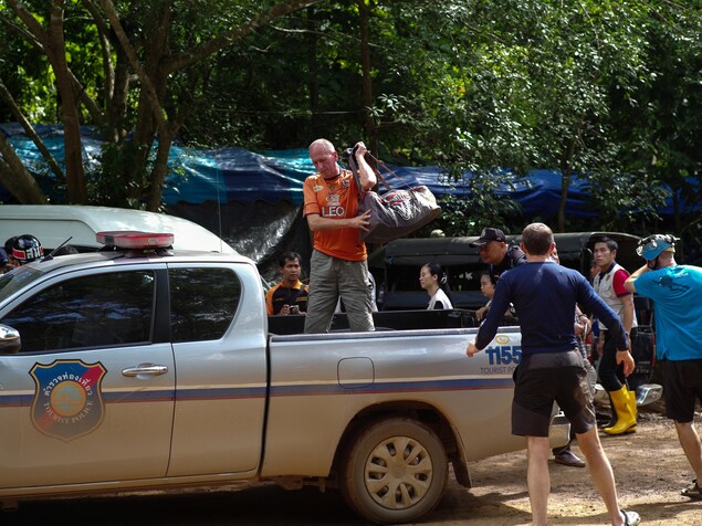 Un homme chauve, qui est debout à l'arrière d'un camion, jette un sac de sport sur son épaule gauche. D'autres personnes se trouvent près du camion. Un homme chauve, qui est debout à l'arrière d'un camion, jette un sac de sport sur son épaule gauche. D'autres personnes se trouvent près du camion.
