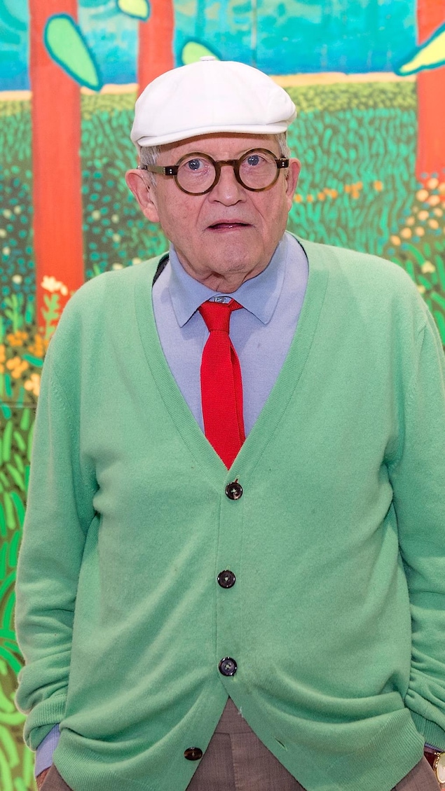 Portant un béret et des lunettes rondes, David Hockney a les mains dans ses poches et regarde l'objectif devant une de ses toiles.