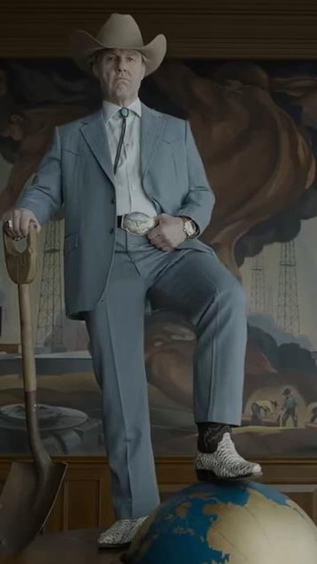Tom Cruise se tient debout sur un bureau. Il tient une pelle dans sa main droite et il y a une balle à l'image de la Terre sous son pied gauche,