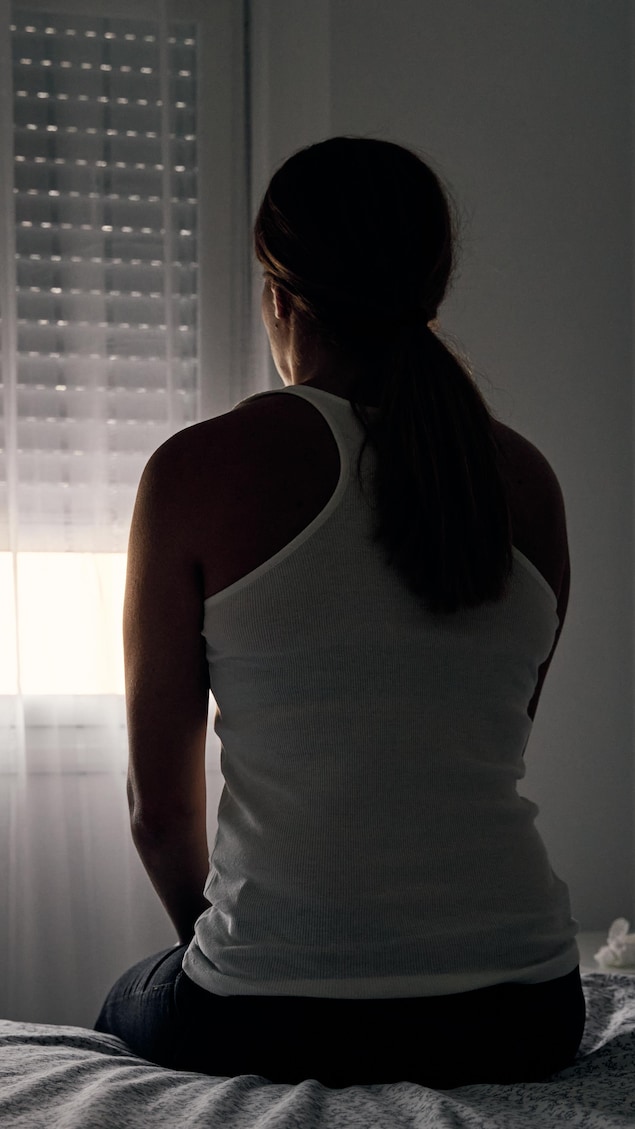 Une femme, assise sur son lit, regarde par la fenêtre.