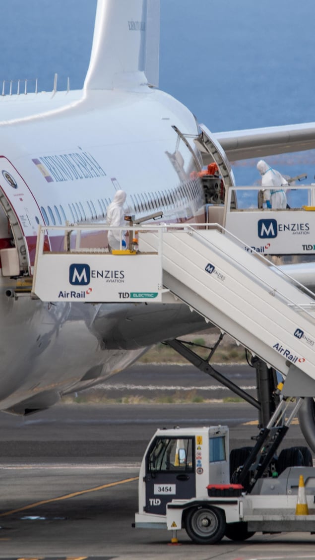 Des personnes vêtues de combinaisons de protection se tiennent aux alentours d'un avion sur le tarmac.