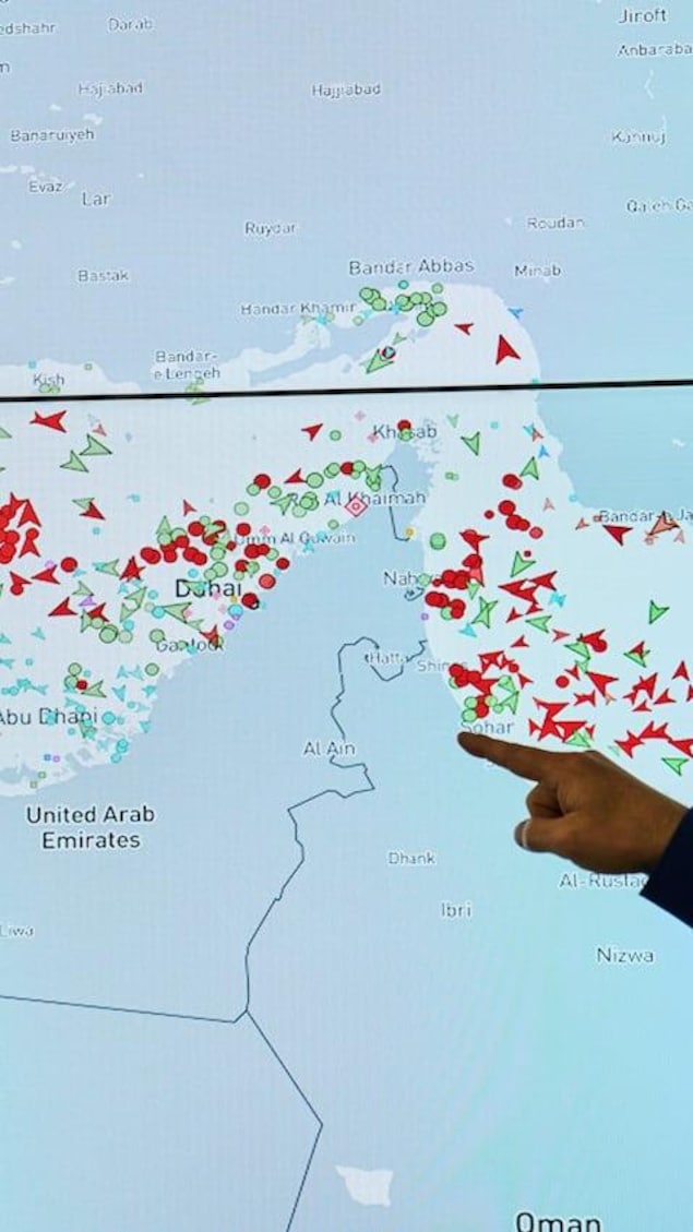 A man looks at a screen displaying a map of the Strait of Hormuz with various dots and arrows representing vessels as someone points to a specific location.