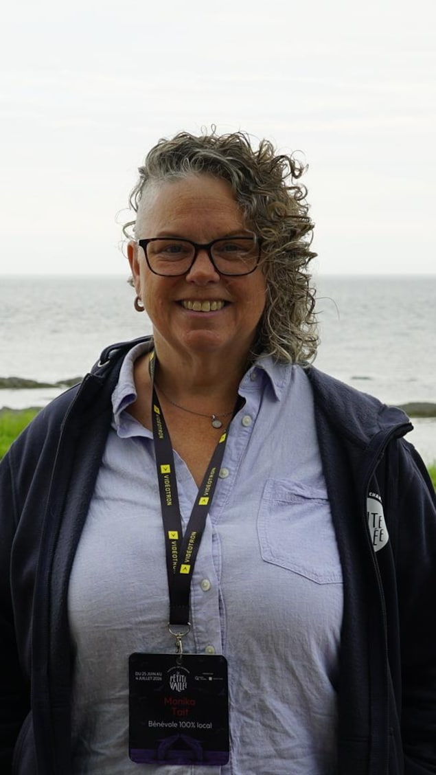 La mairesse porte des lunettes et a des cheveux bouclés. Elle se tient devant une vue dégagée sur le golfe du Saint-Laurent.