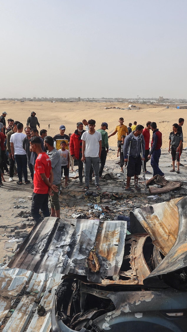 De jeunes hommes et des enfants se tiennent debout au milieu de baraques effondrées sur un vaste terrain sablonneux.