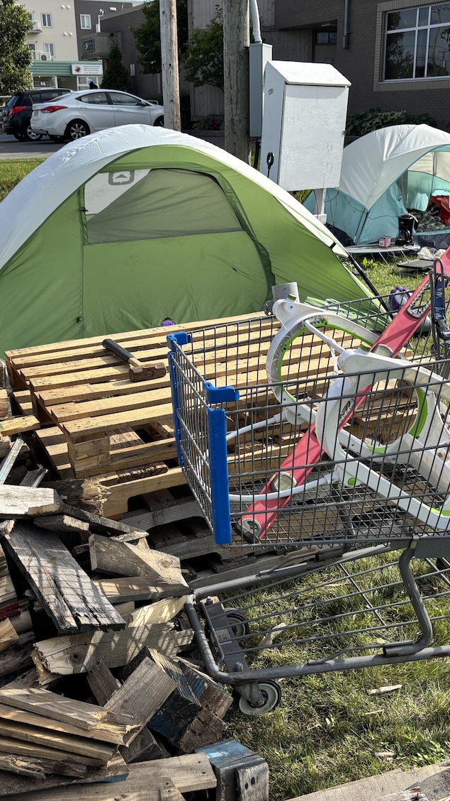 Des tentes de camping sont installées près d'un chariot d'épicerie et d'un empilement de palettes de bois, à Sept-Îles.