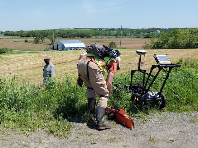 Deux hommes se préparent à fouiller sous le sol dans un champ au moyen d'équipement monté sur un chariot.