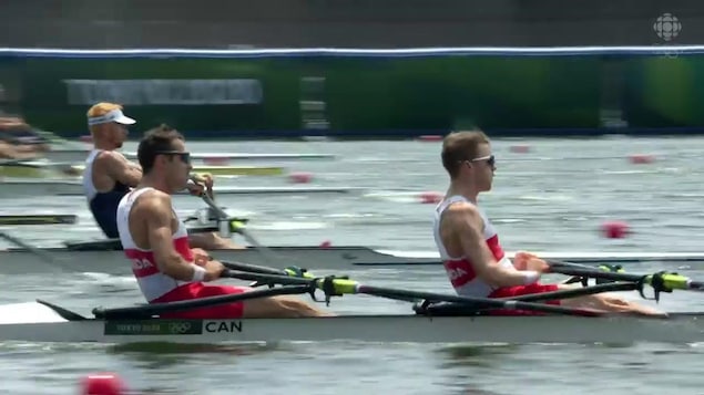 Deux hommes rament avec puissance sur un aviron. Un homme avec une casquette et les cheveux blonds effectue la même chose en arrière-plan