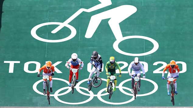 Six athlètes de BMX saute sur une première bosse après la descente de départ lors d'une épreuve olympique. Le logo géant de la discipline avec l'inscription Tokyo 2020 apparaît en arrière.