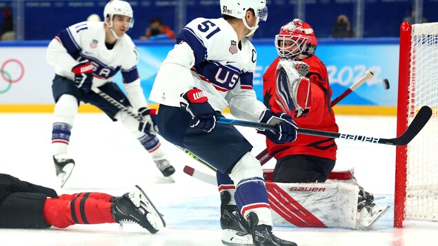 Deux joueurs de hockey en blanc regardent la rondelle pénétrer dans le filet derrière un gardien en rouge pendant un match.