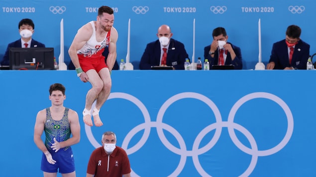 Benjamin Gischard de l'équipe de la Suisse est en plein saut dans les airs durant le temps de pratique pour la routine au sol tandis que le gymnaste brésilien le regarde.