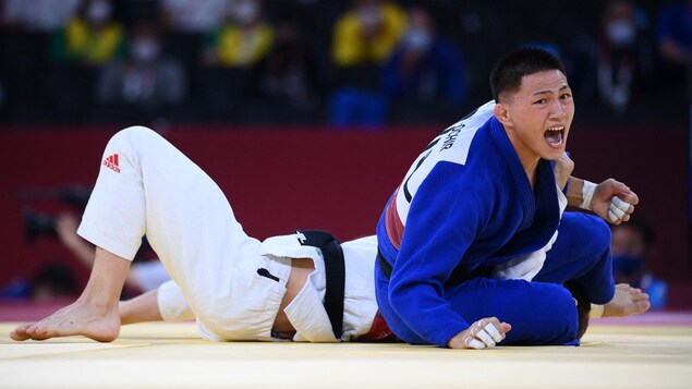 Tsogtbaatar Tsend-Ochir réagit à sa victoire contre le judoka canadien Arthur Margelidon tandis que le judoka canadien est couché au sol derrière le judoka mongole qui est assis sur le côté en train de célébrer.