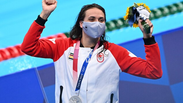 Elle lève les bras en l'air. Elle a sa médaille d'argent au cou et tient un bouquet de fleurs dans sa main gauche