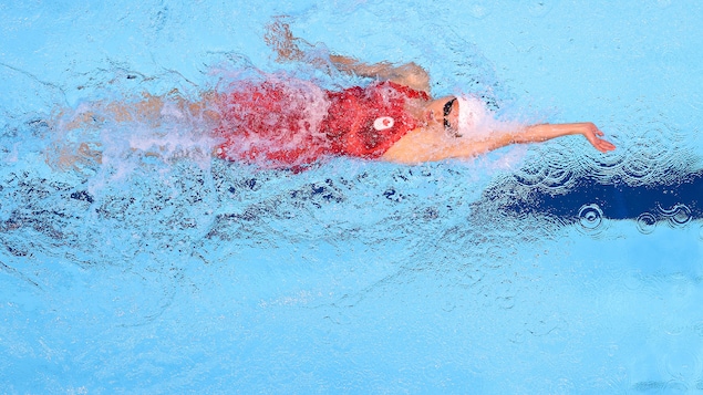 Une jeune athlète nage sur le dos lors d'une course de natation. Elle porte un uniforme aux couleurs du Canada et un casque de bain blanc. 