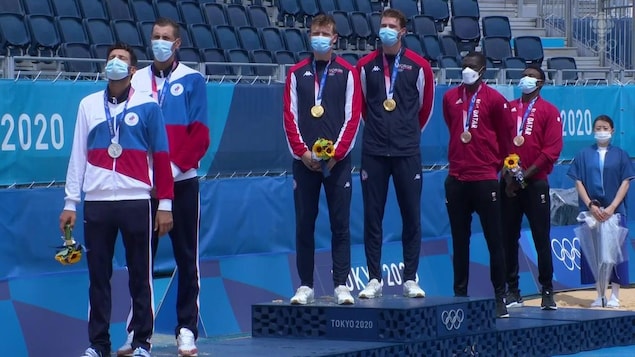 Les trois duos masculins en volleyball de plage se retrouvent sur le podium, tous tournés vers la droite avec leur masque, leur médaille à leur cou et un bouquet de fleurs, les yeux fermés.