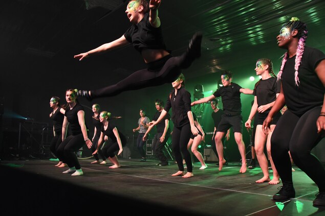 De jeunes danseurs sur la scène habillés collants et teeshirts noirs. Une jeune femme au premier plan saute en faisant le grand écart dans les airs. De jeunes danseurs sur la scène habillés collants et teeshirts noirs. Une jeune femme au premier plan saute en faisant le grand écart dans les airs.