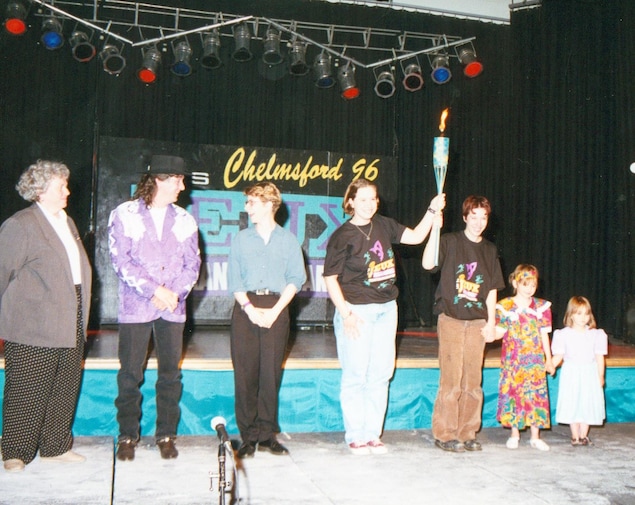 7 personnes devant la scène des troisièmes Jeux franco-ontariens. De gauche à droite, une dame avec pantalons à pois et veston gris. À côté, le chanteur Jean-Guy Chuck Labelle. À côté de lui, une dame avec pantalons noirs et blouse bleue. Au centre, deux jeunes tiennent un flambeau à bout de bras. Finalement à côté d'eux à droite, deux petites filles vêtues de robes. 7 personnes devant la scène des troisièmes Jeux franco-ontariens. De gauche à droite, une dame avec pantalons à pois et veston gris. À côté, le chanteur Jean-Guy Chuck Labelle. À côté de lui, une dame avec pantalons noirs et blouse bleue. Au centre, deux jeunes tiennent un flambeau à bout de bras. Finalement à côté d'eux à droite, deux petites filles vêtues de robes.