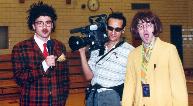 À gauche, un jeune homme hirsute un micro à la main portant un veston à carreaux rouge et noir. Au centre un technicien avec une caméra de télévision sur l'épaule. À droite, un jeune homme vêtu de beige et de jaune feint la surprise devant le photographe. À gauche, un jeune homme hirsute un micro à la main portant un veston à carreaux rouge et noir. Au centre un technicien avec une caméra de télévision sur l'épaule. À droite, un jeune homme vêtu de beige et de jaune feint la surprise devant le photographe.