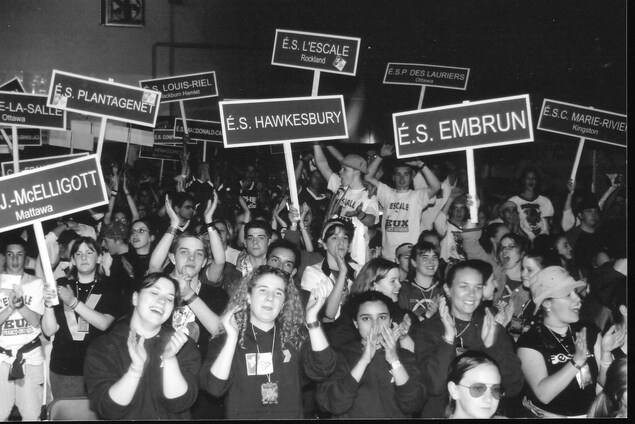 Photo en noir et blanc d'un grand groupe souriant et applaudissant. 9 pancartes sont visibles. Photo en noir et blanc d'un grand groupe souriant et applaudissant. 9 pancartes sont visibles.